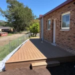 Installation of a new composite deck with a diagonal pattern next to a brick house under a clear blue sky.
