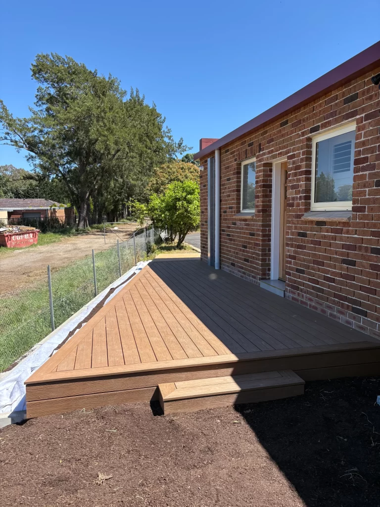 Installation of a new composite deck with a diagonal pattern next to a brick house under a clear blue sky.