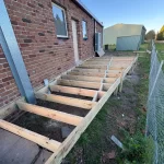 Installation of a new treated timber deck frame attached to a brick house on a clear, sunny day.