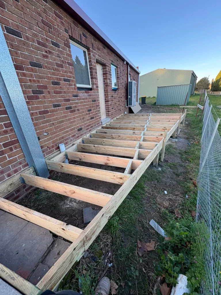 Installation of a new treated timber deck frame attached to a brick house on a clear, sunny day.
