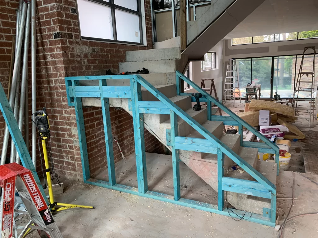 Installation of a blue-painted timber frame alongside a new concrete staircase, within a residential construction site.