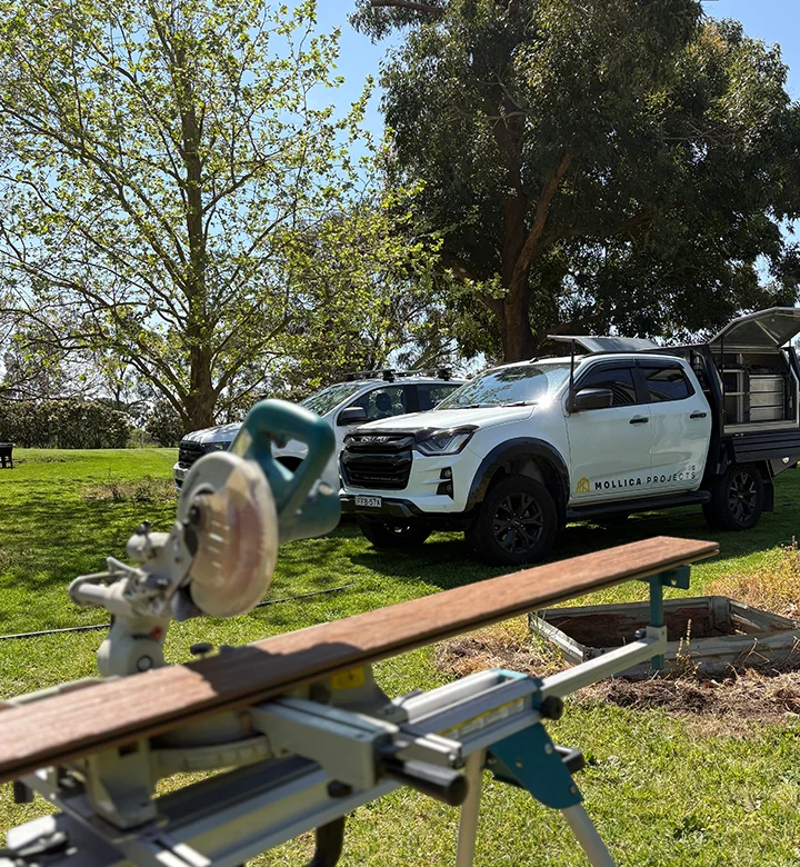 Carpenter working on a decking project with professional power tools and work vehicles nearby.